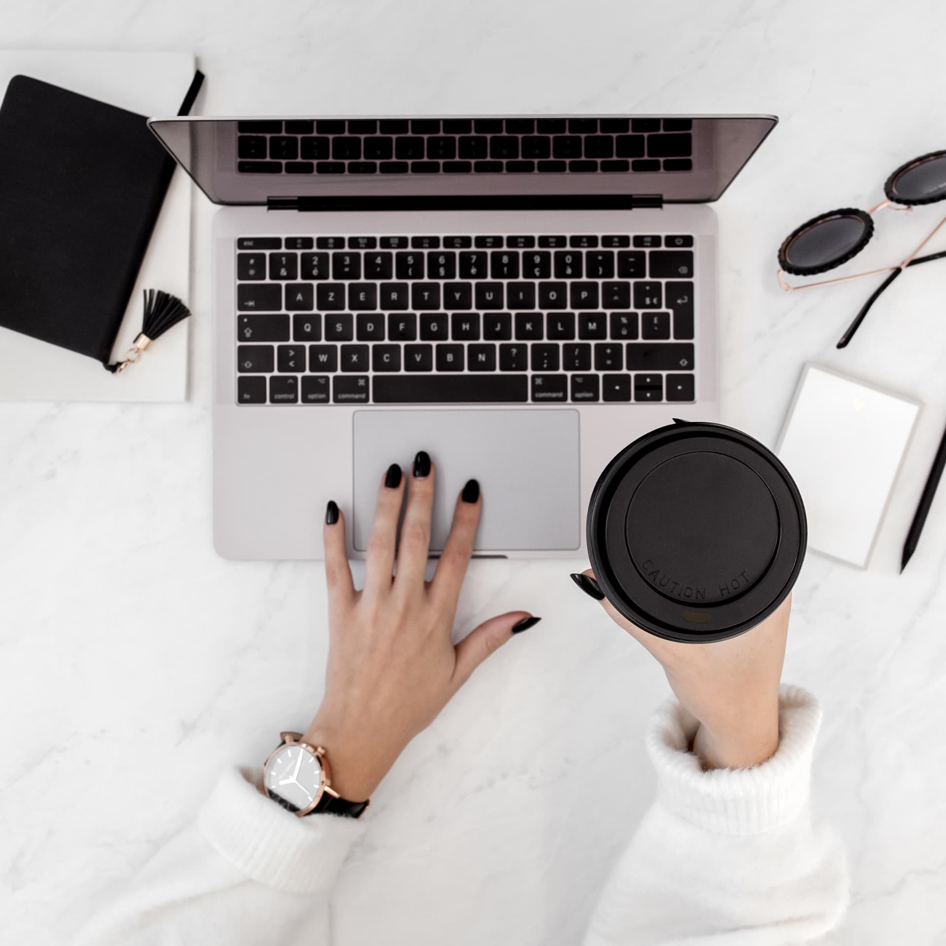Hands working at a laptop beside a black coffee cup on a marble desk.