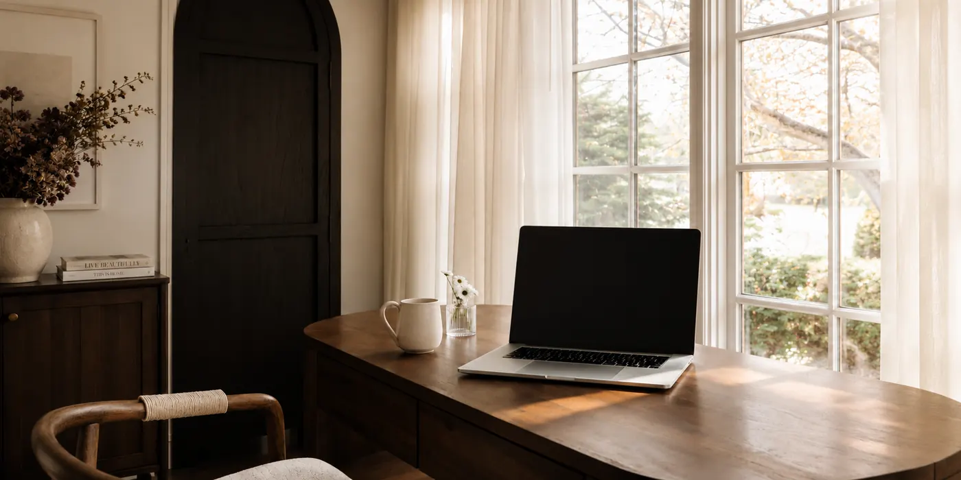 A laptop and coffee cup on a wooden desk beside tall windows.