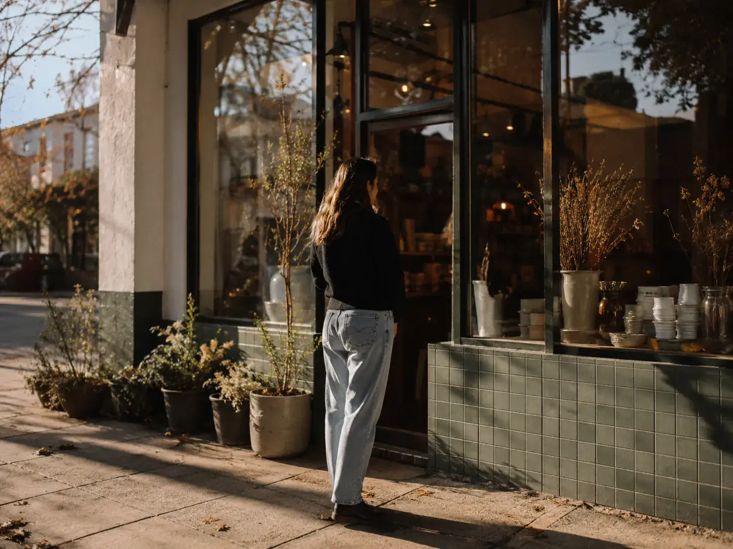 A woman standing on a sunlit sidewalk looking into a shop window.