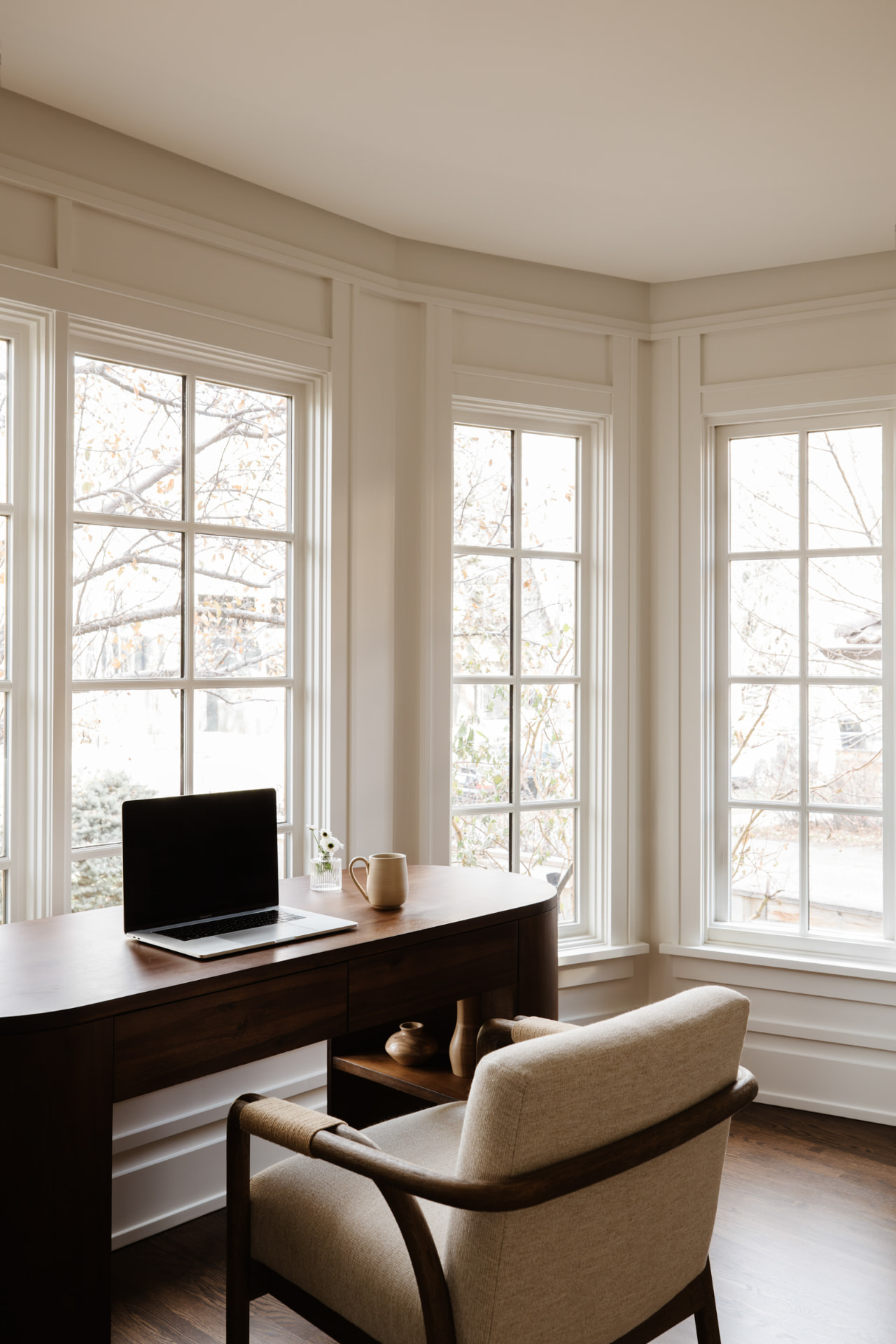 A bright bay-window office with a dark wood desk and chair.
