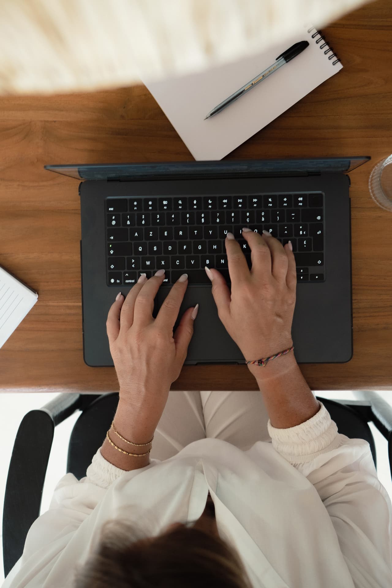 Hands typing on a laptop at a wooden desk with a notebook and pen.